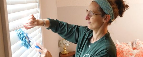 A woman dusts blinds in a bedroom.